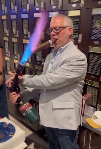 A person using a large industrial blowtorch to light a small cigar.